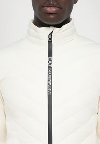 Šviesiai smėlio spalvos izoliuota striukė su aukštu apykakle, siuvinėtu raštu ir juodu užtrauktuku, ant kurio yra "EA7 Emporio Armani" ženklas.