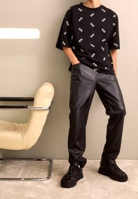 Camiseta negra con un patrón del logo "GODS" en blanco, combinada con pantalones de cuero negros con un detalle de cremallera. Zapatillas negras completan el conjunto.