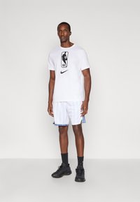 Tricou alb Nike cu logo-ul NBA, asortat cu o pereche de pantaloni scurți în dungi albastre și albe. Pantofi sport negri și șosete completează ținuta.