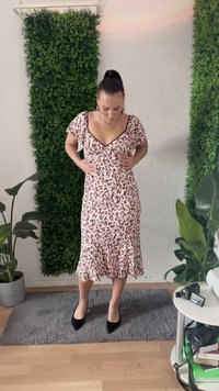 Vestido midi floral em rosa claro com detalhes em preto e vermelho. Apresenta mangas curtas e soltas, decote em V e uma bainha com babados. Sapatos pretos de bico fino.