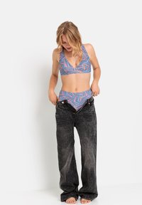 Komplet kopalk, ki vključuje modro in roza bikinije z trapezastim vzorcem, v kombinaciji z ohlapnimi črnimi svetlimi kavbojkami, stoji na bosih nogah.