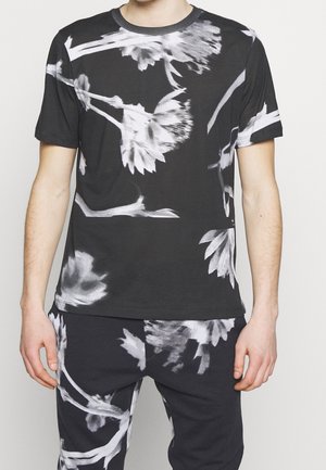 Czarna koszula z krótkim rękawem i spodnie z dużym białym kwiatowym wzorem, noszone przez stojącą osobę, ramiona luźno opuszczone przy bokach.