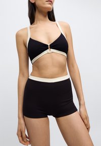 Svart ribbet bikinioverdel med kremfarget kant og knutedetalj i midten, kombinert med høytlivs svarte ribbede shorts. Jevn tekstur over hele plagget.