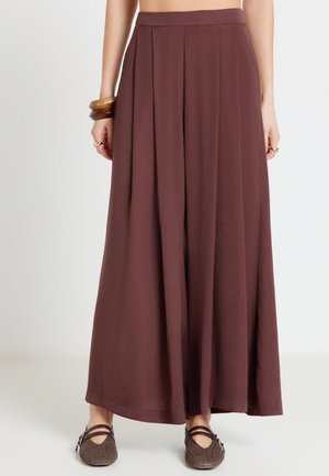 Pantaloni marroni a gamba larga con pieghe indossati con scarpe Mary Jane marroni intrecciate e braccialetti di legno al polso della modella.