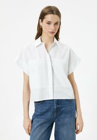 Camicia bianca a maniche corte con colletto e taschini frontali, realizzata in tessuto liscio. Abbinata a jeans di denim blu, che mostrano uno stile informale.