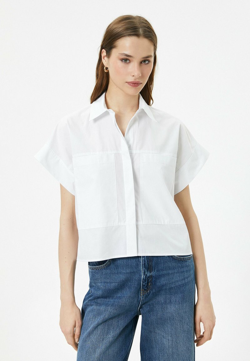 Camicia bianca a maniche corte con colletto e taschini frontali, realizzata in tessuto liscio. Abbinata a jeans di denim blu, che mostrano uno stile informale.