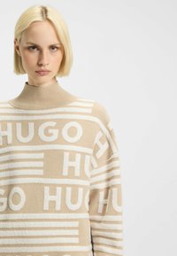 Beige neule korkealla kauluksella, jossa on valkoisia raidallisia kuvioita ja "HUGO"-teksti etupuolella. Pehmeä neulos, rento malli.