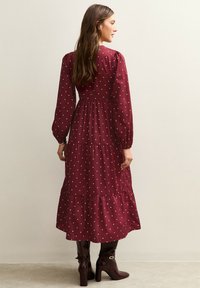 Langes burgunderrotes Kleid mit weißen Polka-Dots, Puffärmeln, gestuftem Rock und tailliertem Schnitt. Kombiniert mit knielangen braunen Stiefeln.