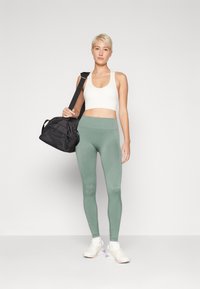 Bijela sportska brusa, mint-zelene tajice s rebrastom teksturom i bijele atletske cipele. Osoba drži crnu teretanu torbu s remenom za rame.