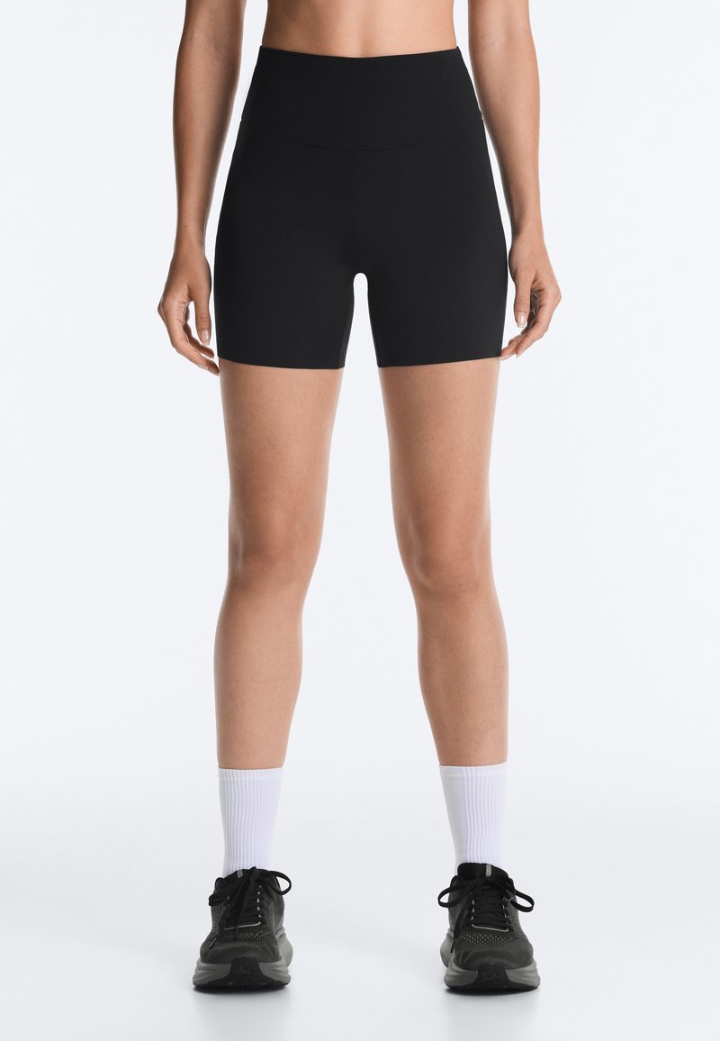 Femme portant un short noir taille haute ajusté, des chaussettes blanches montantes et des chaussures de sport noires, debout sur un fond blanc.