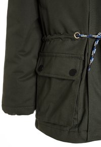 Veste olive en tissu texturé, avec une taille ajustable par cordon, une poche à bouton-pression et un ourlet réglable avec des accents de cordon contrastants.