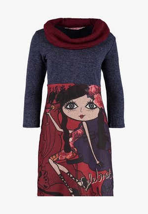Femme marchant dans une robe bleu foncé avec un imprimé coloré d'une fille de dessin animé, des collants noirs, des bottines noires, et portant un grand sac à bandoulière en suède beige.