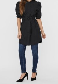 Schwarzes Baumwollmischung-Kleid mit Puffärmeln, Knopfverschluss und tailliertem Bund. Kombiniert mit schmalen blauen Jeans und schwarzen Absätzen.