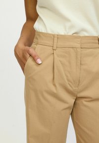 Bež hlače visokog struka napravljene od mješavine pamuka. Ima bočne džepove, pliseje sprijeda i glatku, mekanu teksturu.