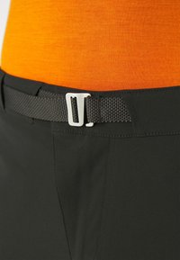 Mustat housut, joissa on kiinteä tekstuuri, varustettuna kudotulla vyöllä ja hopeavöillä. Alta näkyy osittain oranssi paita.