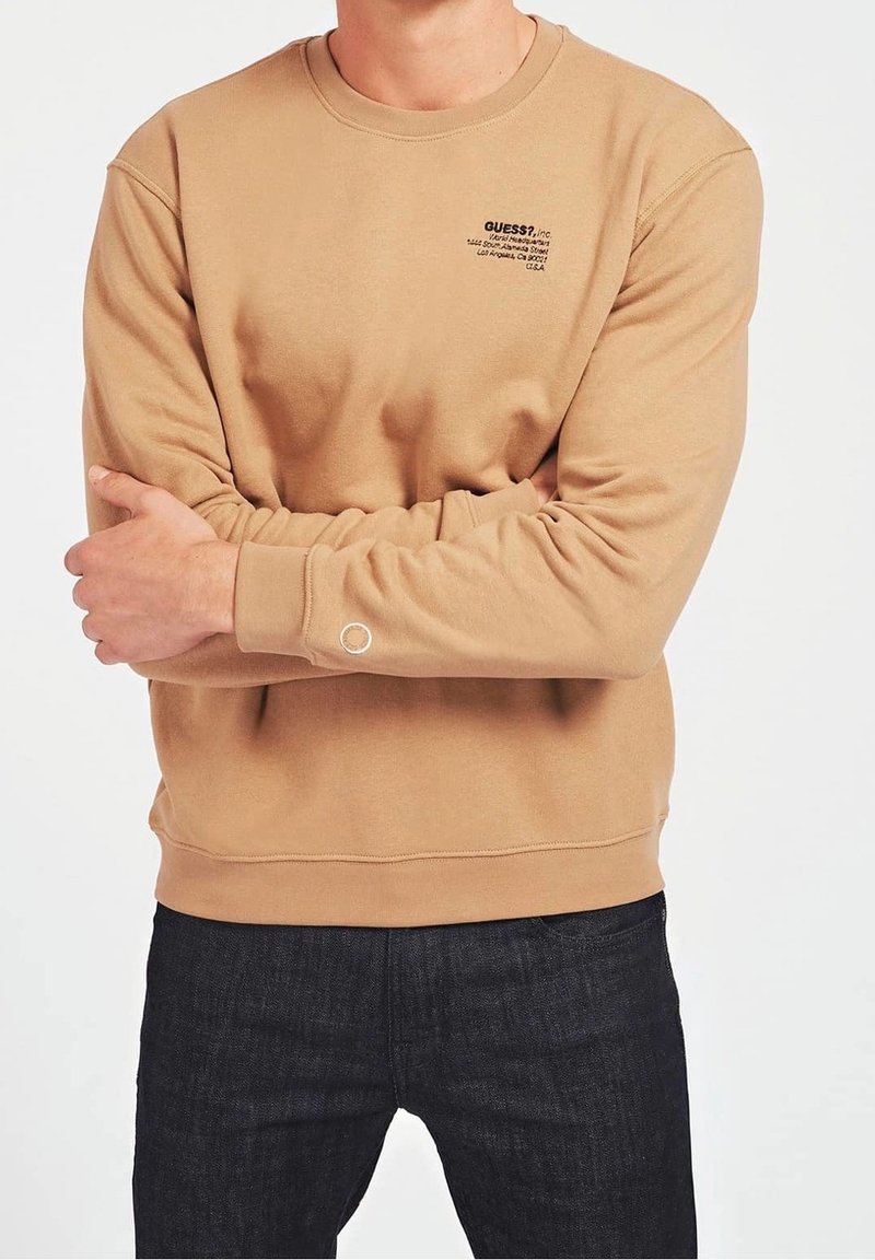 Felpa in cotone beige con collo tondo, polsini a costine e un piccolo logo nero sul petto. Indossata con jeans scuri.