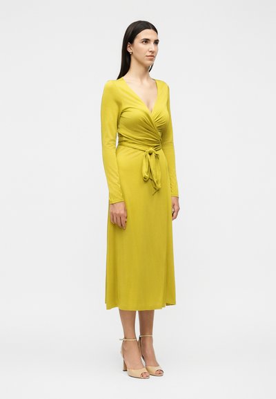 Frau in einem gelben Wickelkleid mit langen Ärmeln, in der Taille gebunden, stehend mit neutralem Gesichtsausdruck, beige offene Pumps vor weißem Hintergrund.