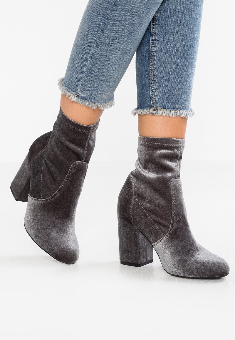 Femme portant des bottines grises en velours avec talons carrés et jean bleu à ourlet effiloché, debout sur une surface blanche.