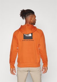 Felpa arancione con cappuccio, realizzata in tessuto morbido. Presenta una patch logo nera sulla parte superiore della schiena. Design semplice e sportivo.