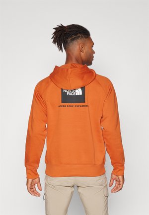 Felpa arancione con cappuccio, realizzata in tessuto morbido. Presenta una patch logo nera sulla parte superiore della schiena. Design semplice e sportivo.