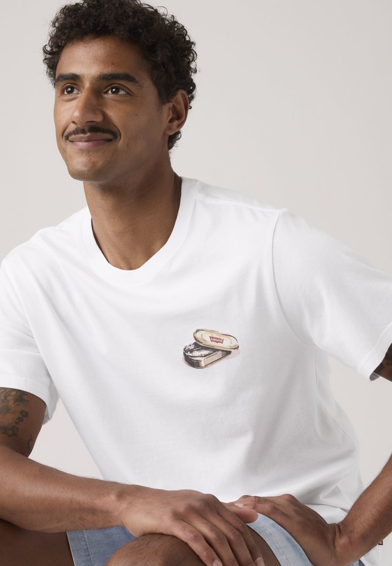 Man met krullend haar en snor, draagt een wit T-shirt met een kleine afbeelding van een open blikje op de borst, zittend met handen rustend op de knie.
