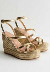 Sandalias de cuña metálica dorada con diseño de tira cruzada, detalles de yute trenzado en el tacón y una plantilla rosa suave.