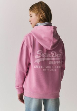 Pink hooded sweatshirt made of soft fabric, featuring white graphics on the back, and ribbed cuffs and hem. Worn with light denim.