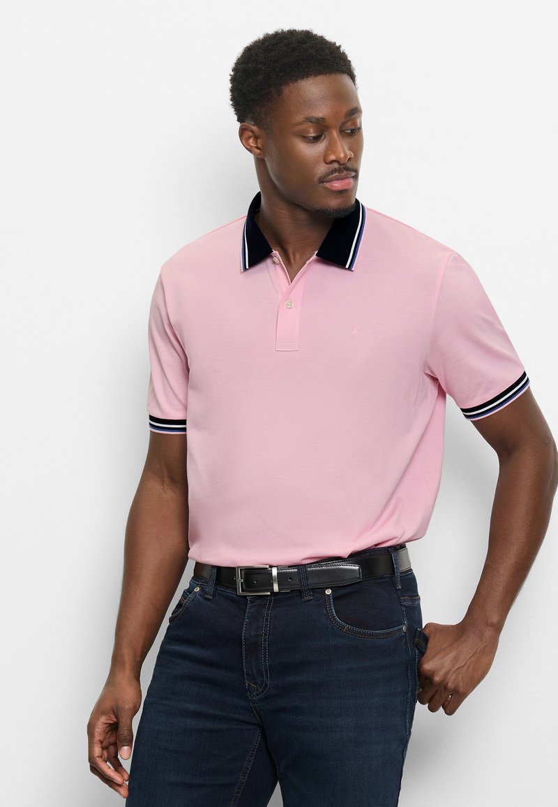 Mann trägt ein rosa Polohemd mit schwarzem Kragen und Ärmelsaum, dunkle Jeans und schwarzen Gürtel, schaut nach links.