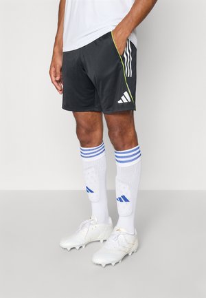 Shorts de sport noirs avec des accents latéraux verts, chaussettes blanches avec des rayures bleues et chaussures de soccer blanches. Tous les articles comportent des logos Adidas.
