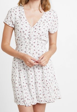 Une femme porte une robe courte blanche à fleurs avec des boutons et des manches courtes, les mains doucement croisées à la taille, sur un fond blanc uni.