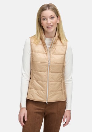 Beige Steppweste mit gesteppten Abschnitten, durchgehendem Reißverschluss und Stehkragen, getragen über einem weißen Langarmoberteil. Braune Wildleder-ähnliche Hosen.