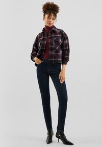 Károvaná bunda v purpurovej a čiernej farbe s gombíkmi, nosená cez bordový rolák a tmavé úzke rifle. Oblečenie dopĺňajú čierne špicaté lodičky.