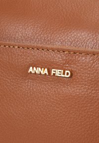 Brown textured leather with gold "ANNA FIELD" logo plate. Features visible stitching and smooth surface. Minimalist design.