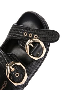 Sandale tissée noire avec deux larges brides, présentant des boucles dorées. La semelle est lisse et plate, et la texture est tressée.