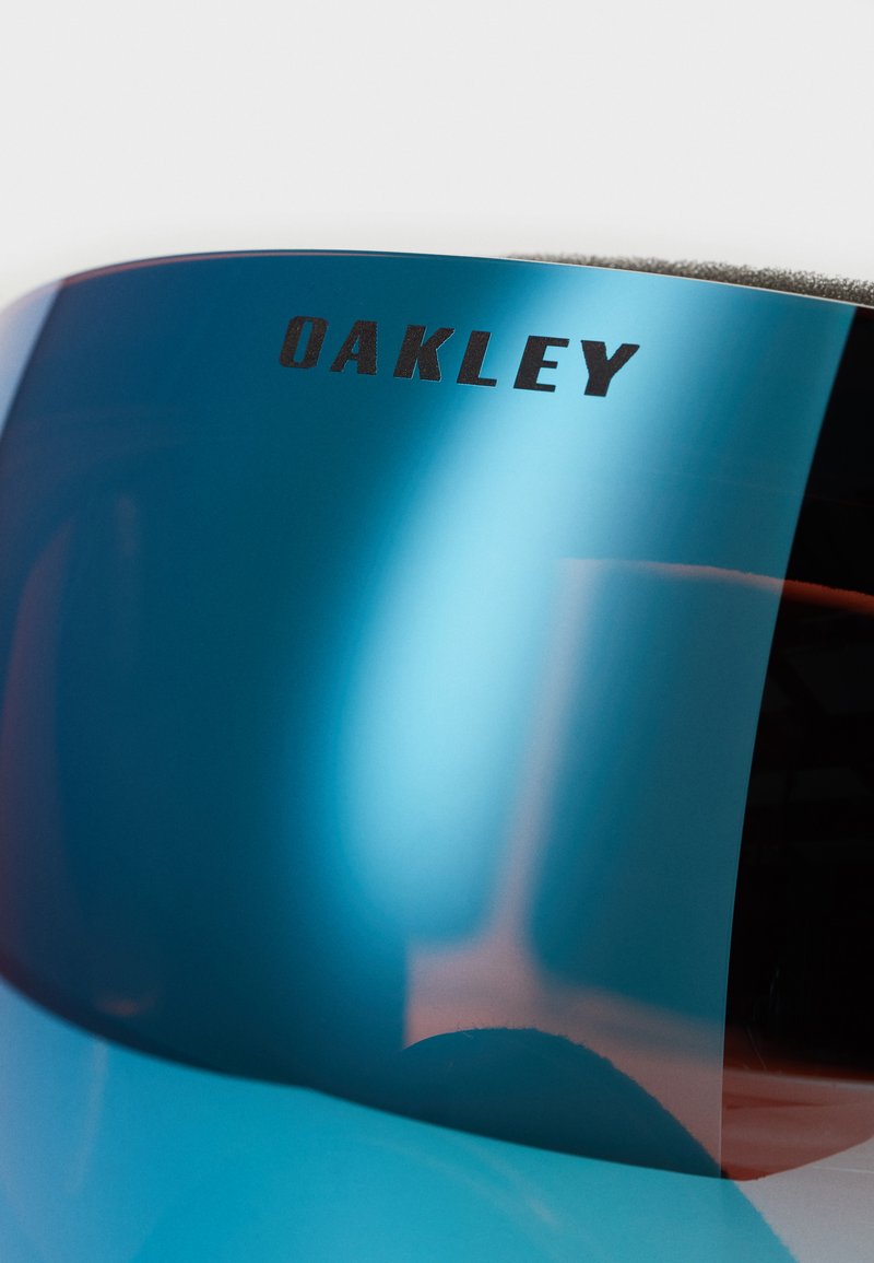 Gafas de sol con tono azul y lentes reflectantes, que presentan un prominente logo de 'Oakley' en negro. Superficie lisa y forma aerodinámica.