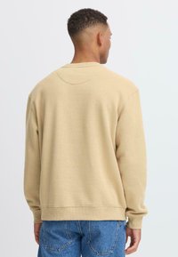 Beige sweatshirt with a round neckline and ribbed cuffs. Features a relaxed fit and smooth texture, paired with faded blue jeans.