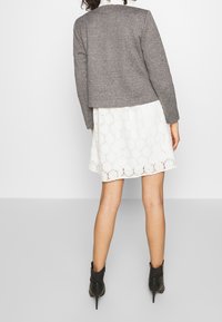 Veste grise texturée portée sur une robe blanche en dentelle avec un motif floral. Des bottines noires à bout pointu complètent la tenue.