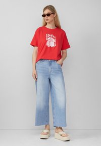 Rode katoenen t-shirt met witte grafische tekst, gecombineerd met lichtblauwe wijde spijkerbroeken en crèmekleurige platte sandalen met bandjes.
