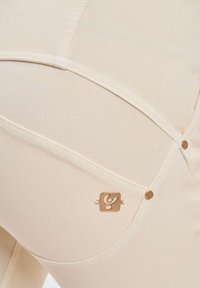 Nærbilde av beige bukser med detaljert søm, messingnitter og et lite rektangulært metallmerke på baklommeområdet.