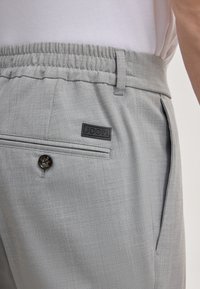 Hellgraue Hose aus strukturiertem Stoff, mit elastischem Bund, Knopfverschluss und einem schwarzen Logopatch. Verfügt über eine Gesäßtasche und seitliche Nähte.