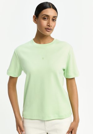 Frau mit zurückgebundenem dunklem Haar, die ein hellgrünes Kurzarmshirt und weiße Hosen vor einem schlichten weißen Hintergrund trägt.