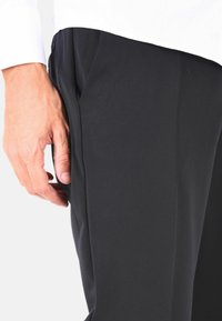 Pantaloni neri a gamba dritta, caratterizzati da una sottile trama rigata. Una mano poggia sulla tasca laterale, evidenziando il design del capo.