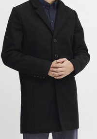 Schwarzer Wollmantel mit figurbetontem Schnitt, gekerbtem Revers, zwei großen Knöpfen und zusätzlichen Knopfdetails an den Manschetten.