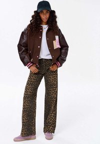 Kobieta z długimi kręconymi włosami, nosząca czarną czapkę, brązową kurtkę typu varsity, białą koszulkę, spodnie w panterkę oraz różowe trampki, stoi z rękami w kieszeniach.