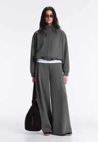 Femme portant une veste zippée gris foncé et un pantalon à jambes larges, tenant un sac noir, avec de longs cheveux foncés et des lunettes de soleil.