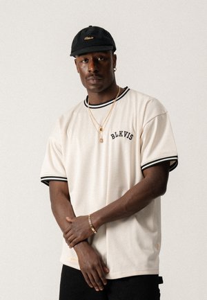 Maillot en mesh crème à manches courtes avec des accents noirs, arborant le logo "BLKVIS" sur la poitrine et un col à côtes. Porté avec un pantalon noir et des accessoires.