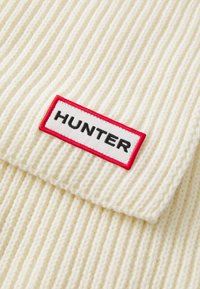 Beanie a maglia color crema con texture a coste e un'etichetta bianca con bordo rosso su cui è scritto "HUNTER" in lettere nere.