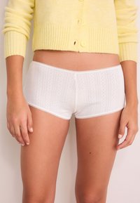 White cotton shorts with a textured pattern and lace trim at the waistband. The model wears a yellow ribbed cardigan.