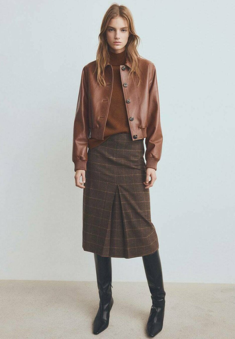 Veste aviateur en cuir marron avec fermeture à boutons sur un pull marron, associée à une jupe midi à carreaux et des bottes noires montantes.
