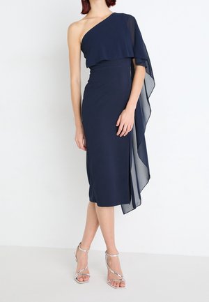 Kobieta ubrana w granatową, midi sukienkę na jedno ramię z przepływającym, przezroczystym kapturem, zestawioną z srebrnymi sandałami na wysokim obcasie.